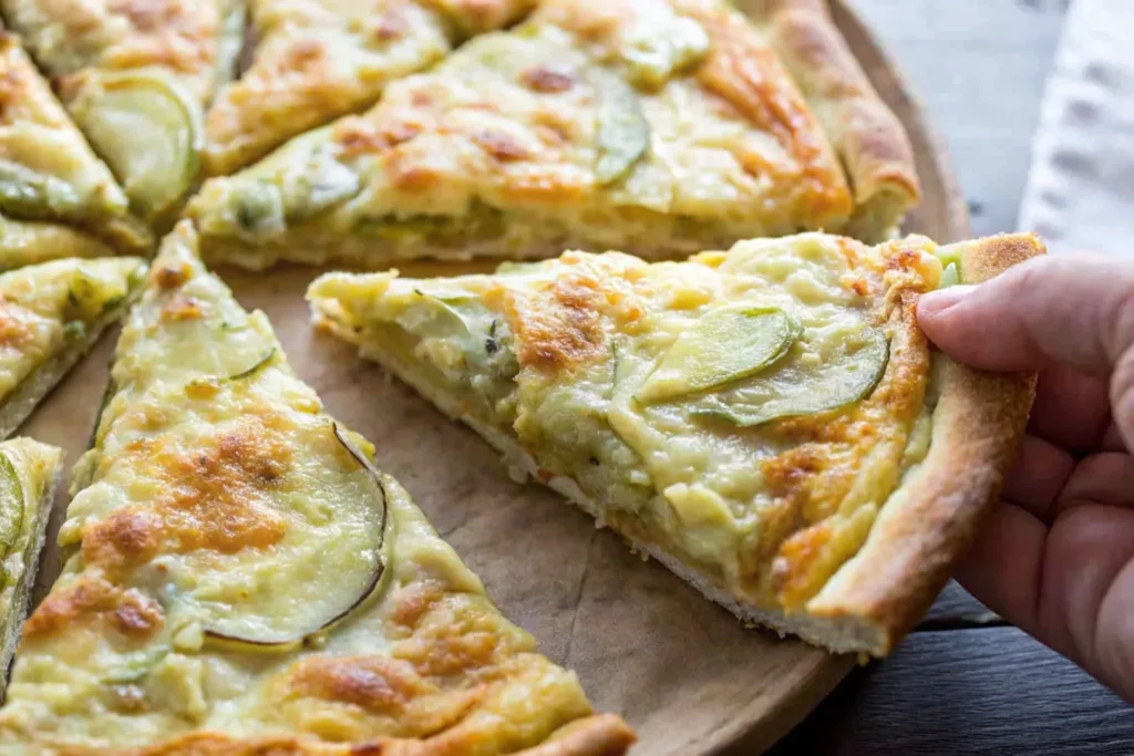 A hand lifting a slice of pickle pizza with melted cheese and sliced pickles, served on a wooden pizza tray.